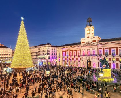 Madrid, Spain - December 18, 2023. Festive crowd gathers around a great golden Christmas tree in the Puerta del Sol square, with the historic Real Casa de Correos Palace illuminated at dusk.